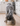 A blue roan cocker spaniel bends down to eat from a beige ceramic spaniel bowl personalized with the name 'Bella' in a bright kitchen with white cabinets and light wood floors.